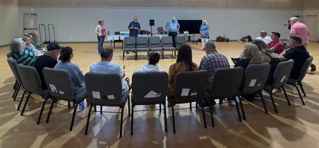 20260711_213924032 - VMTC Community meeting in a gym-like hall with attendees seated in a circle facing four presenters at a long table at the front.