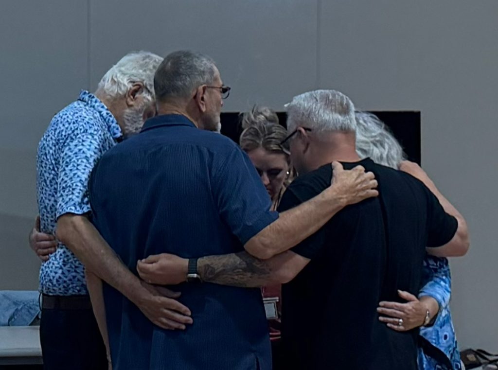 20260711_214218634_iOS - VMTC A group of five adults standing in a tight circle with their arms around each other, offering comfort at an indoor gathering.