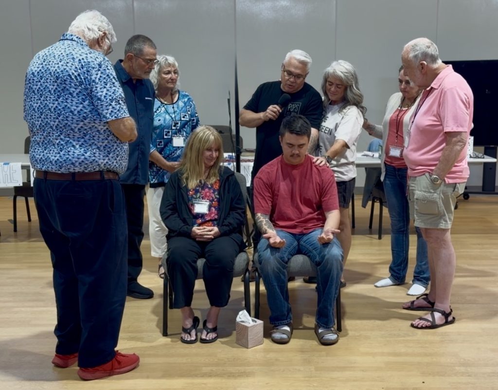 Screenshot - VMTC A group of adults stands around a seated woman and man, placing hands on them in a supportive or blessing ceremony in a meeting room.