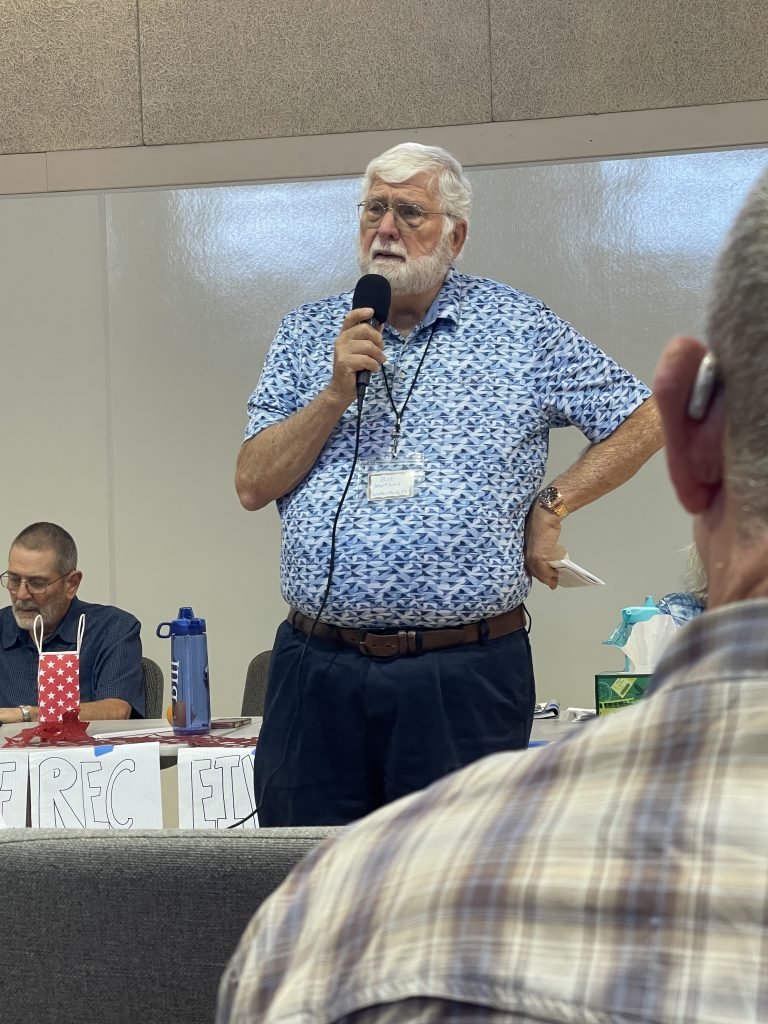 IMG_4667 - VMTC | Victorious Ministry Through Christ Hombre mayor con cabello y barba blancos habla por un micrófono de mano en un panel, vistiendo una camisa azul con patrón y una etiqueta con su nombre en una sala de conferencias.