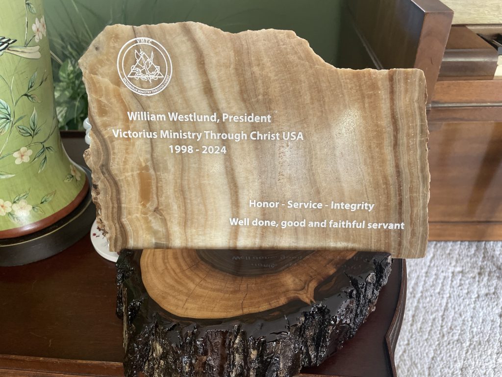 IMG_4184 - VMTC | Victorious Ministry Through Christ Placa conmemorativa de madera con logotipo circular y la inscripción 'William Westlund, President … 1998-2024' en un estante junto a un florero.
